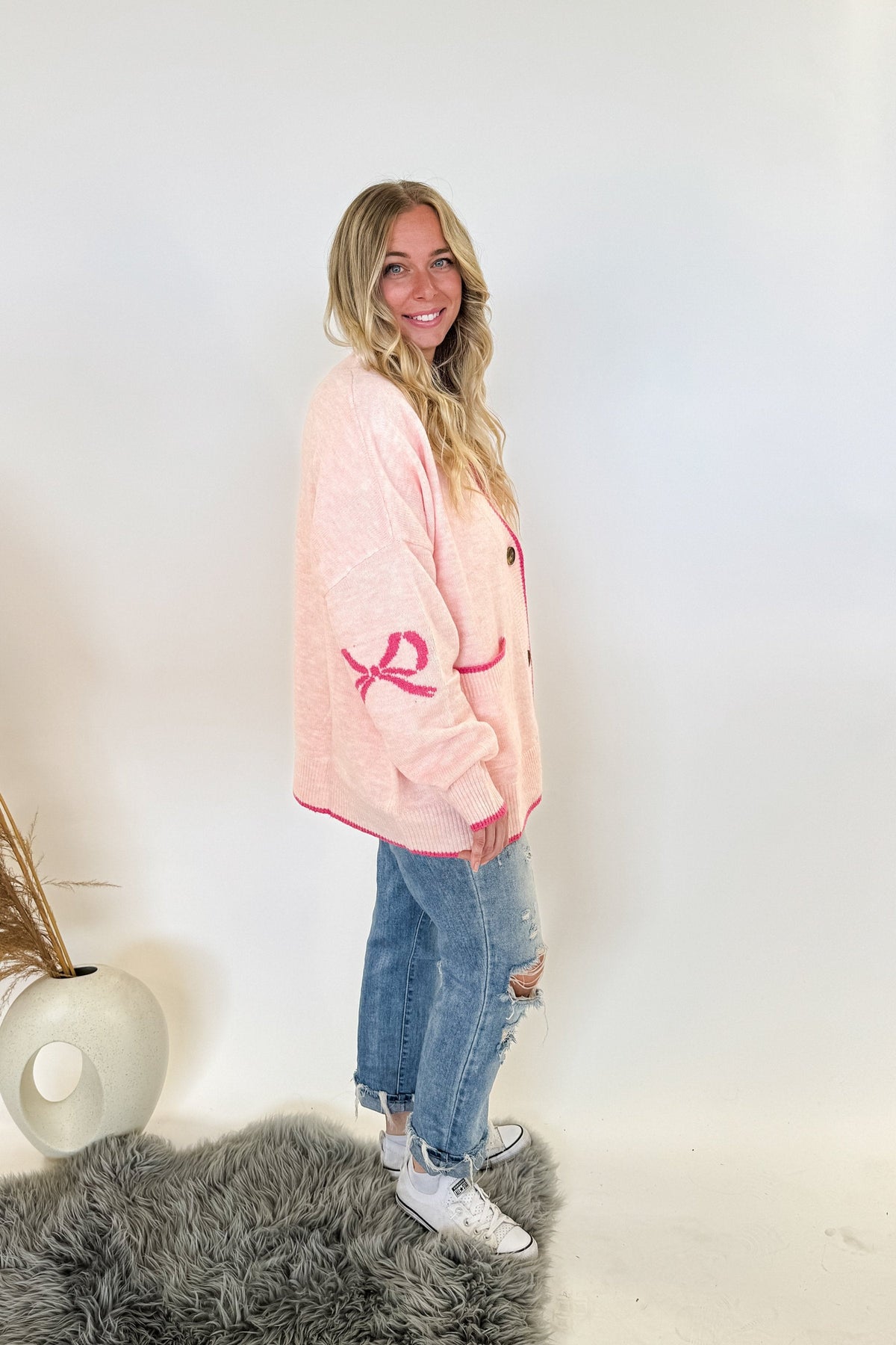 woman stands wearing an oversized cardigan in light pink with bows on the elbows and hot pink trim