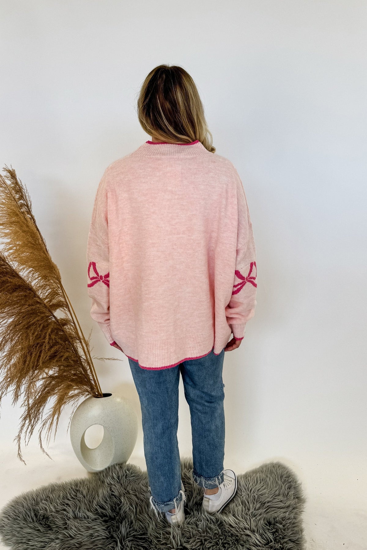 woman stands wearing an oversized cardigan in light pink with bows on the elbows and hot pink trim