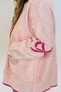 woman stands wearing an oversized cardigan in light pink with bows on the elbows and hot pink trim