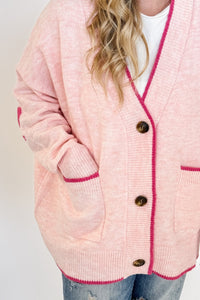 woman stands wearing an oversized cardigan in light pink with bows on the elbows and hot pink trim