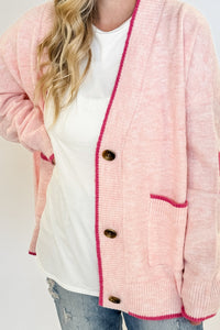 woman stands wearing an oversized cardigan in light pink with bows on the elbows and hot pink trim