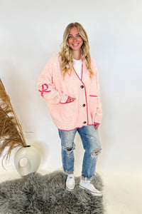 woman stands wearing an oversized cardigan in light pink with bows on the elbows and hot pink trim