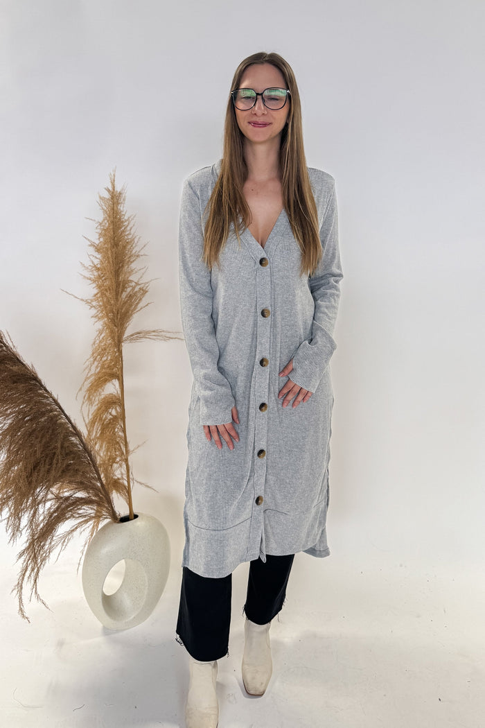 woman wearing very long gray cardigan in a ribbed knit fabric with brown buttons and high slits on the sides. Buttons all the way down the front.