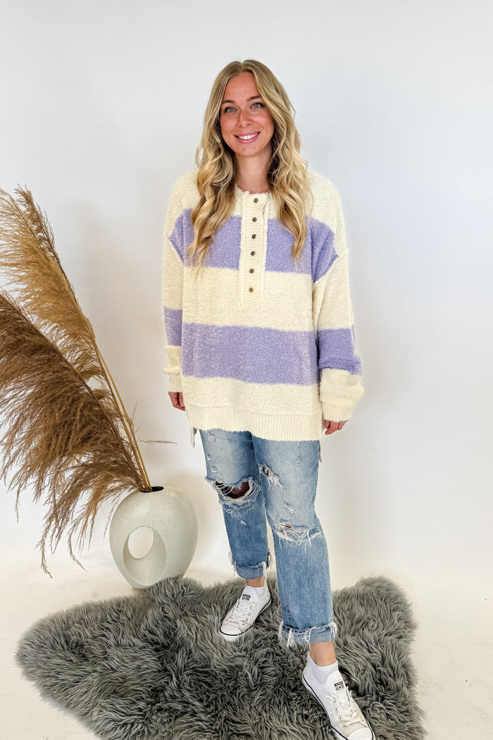 oversize rugby style womans sweater with buttons and purple/ivory stripes. Woman is standing, wearing jeans and sneakers on a white background. 