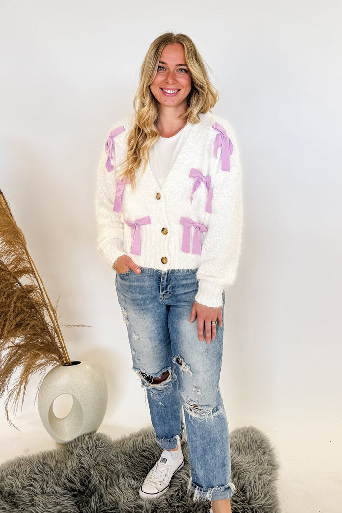 woman stands wearing an ivory sweater with purple ribbons on it 