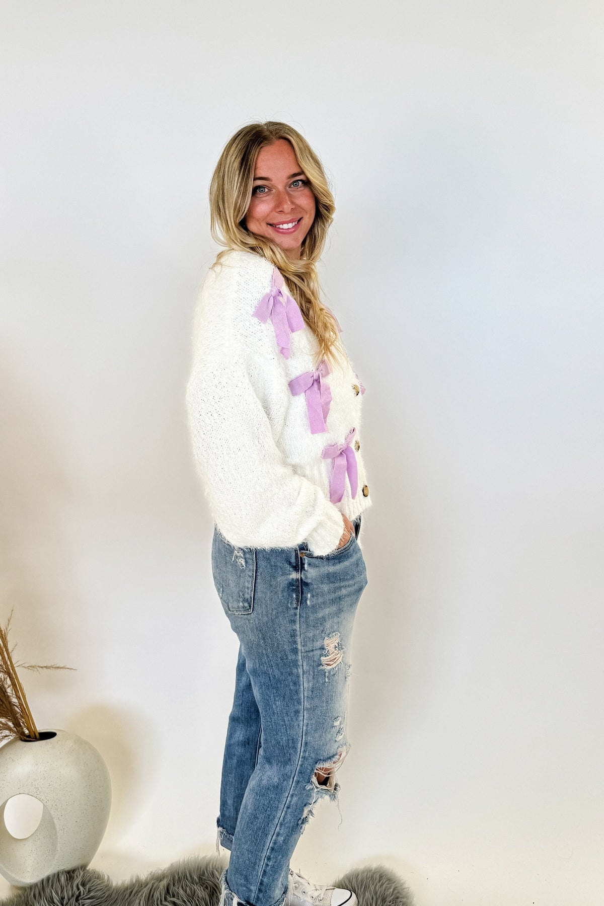 woman stands wearing an ivory sweater with purple ribbons on it 