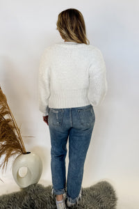 woman stands wearing an ivory sweater with purple ribbons on it 