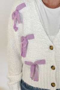 woman stands wearing an ivory sweater with purple ribbons on it 