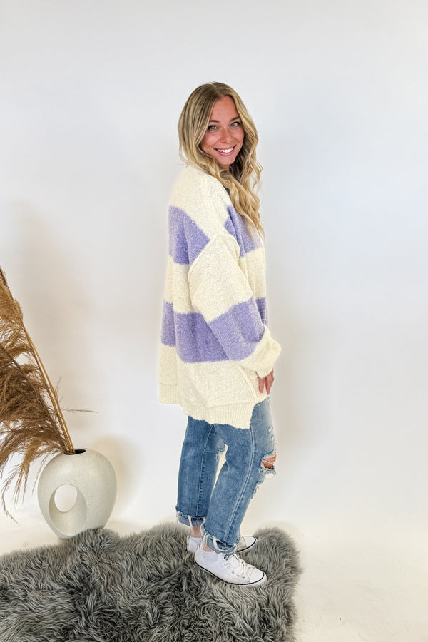 oversize rugby style womans sweater with buttons and purple/ivory stripes. Woman is standing, wearing jeans and sneakers on a white background. 