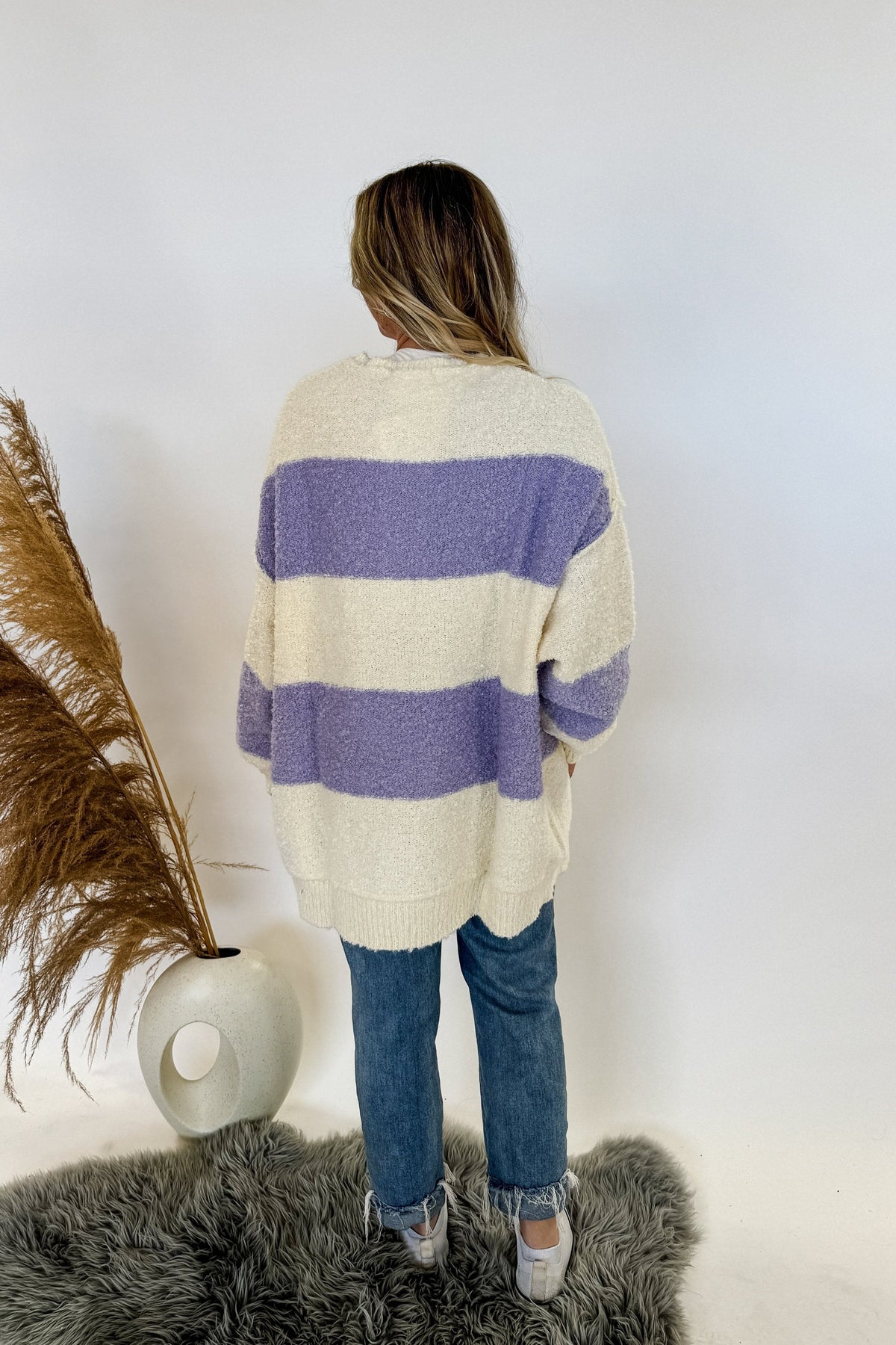 oversize rugby style womans sweater with buttons and purple/ivory stripes. Woman is standing, wearing jeans and sneakers on a white background. 